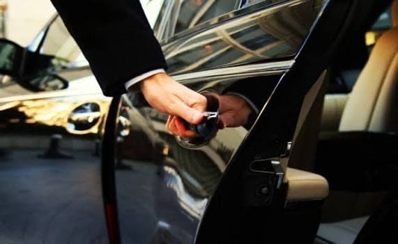 A driver in a dark suit opening the rear door of a black car